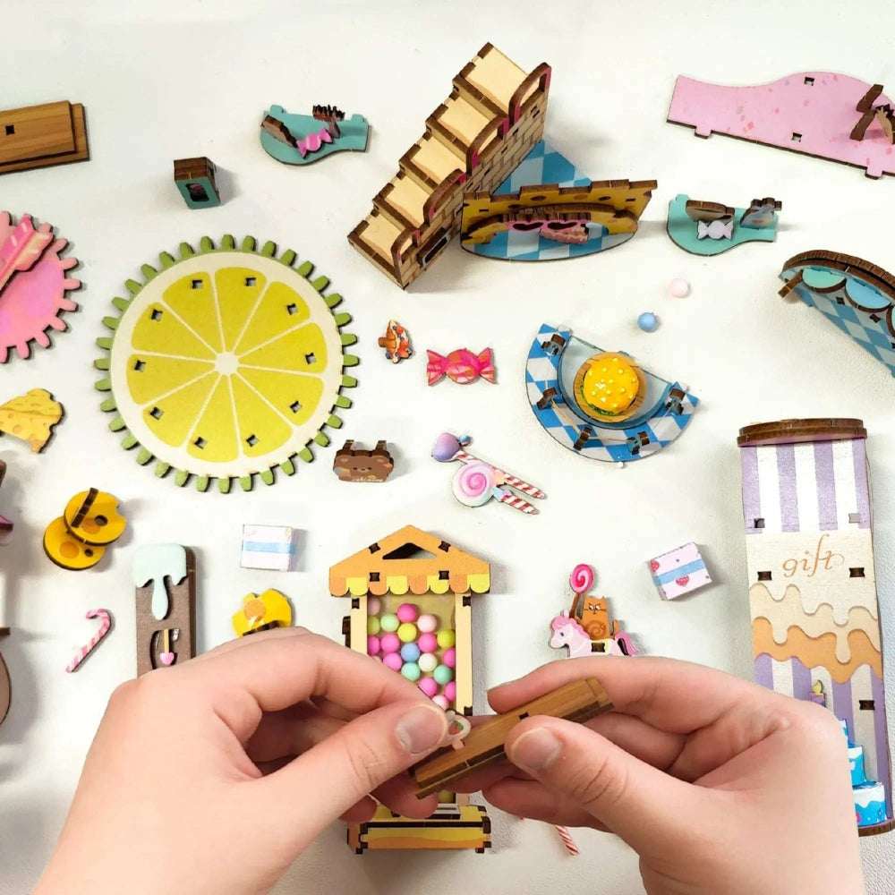 Hands assembling Candy Store miniature pieces beside candy-shaped decorations and a spiral stair.