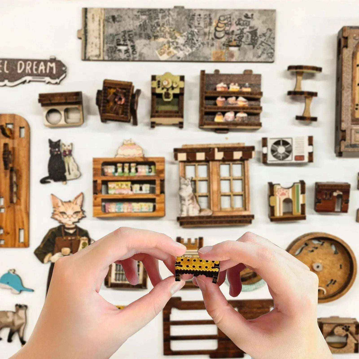 Hands assembling Caramel Dreams miniature parts beside coffee-shop accessories and cat-themed pieces.