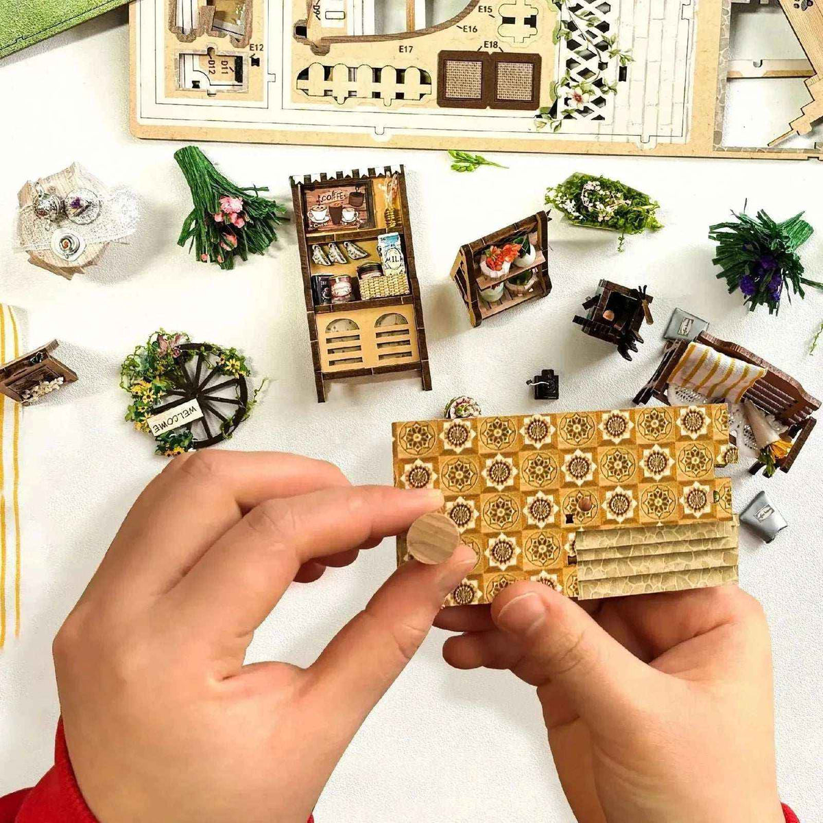 Hands assembling Sylvia's Garden miniature flooring with kit parts arranged on a table.